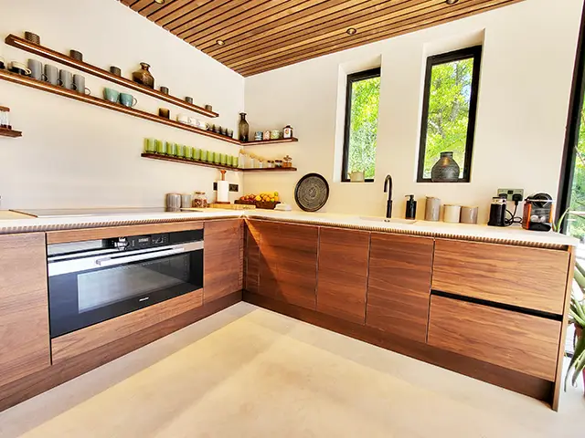 A kitchen with natural wood handleless slab doors, wooden open shelving and an wooden slatted ceiling with downlighters.