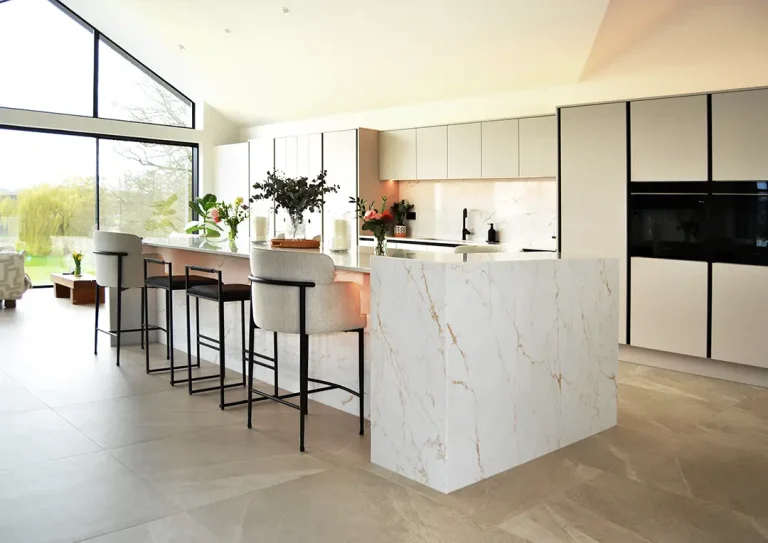Modern open-plan white handleless kitchen with marble patterned quartz island, breakfast bar seating and large picture windows.