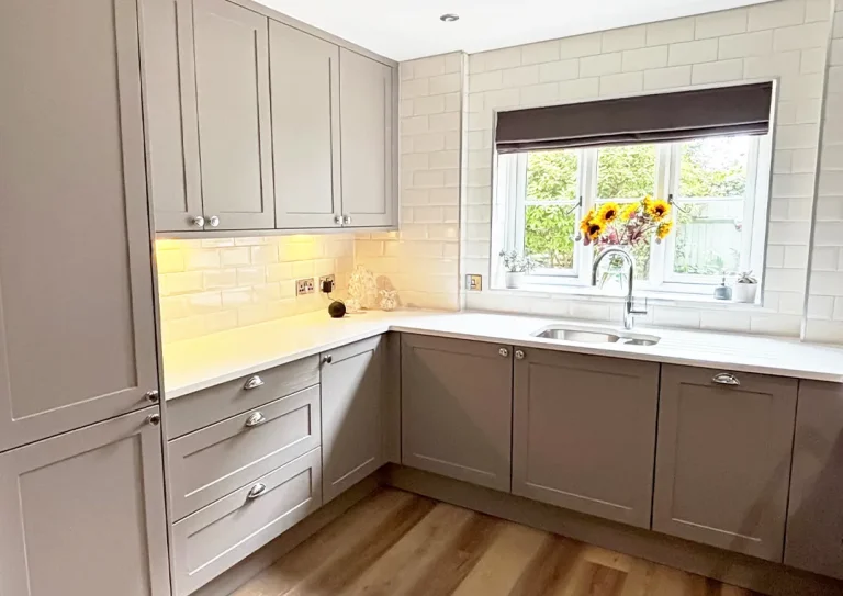 A traditional shaker style kitchen with white metro tiled walls and sunlight shining through a vase of sunflowers sitting on the kitchen window sill.